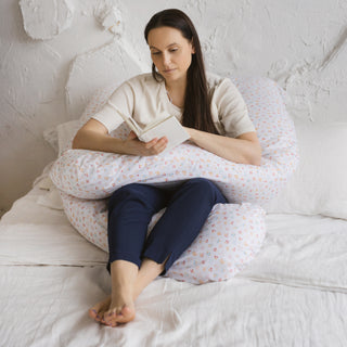 Pregnant woman relaxing with U-shaped maternity pillow in a cozy bedroom setting. By MINICAMP