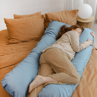 Pregnant woman sleeping on a bed, supported by a MINICAMP U-shaped comfort pillow with a natural sateen cotton cover in blue